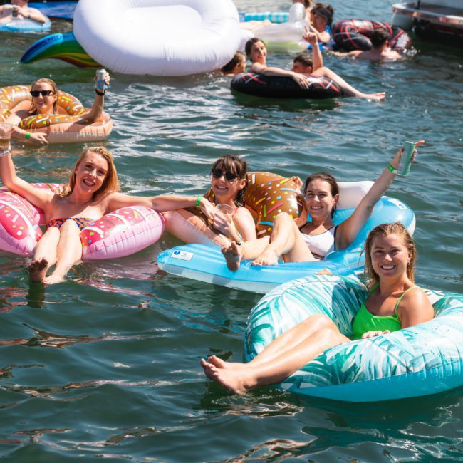 A group of five people floats on inflatable pool rings in a body of water, smiling and holding drinks. Others are seen floating and swimming around them, enjoying what looks like a private yacht charter on Sydney Harbour.