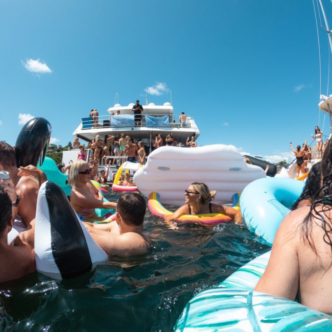 People are gathered in and around a pool with various floats near multiple boats on a sunny day. Some enjoy the water while others relax on boats, making it a perfect scene for a Catamaran party Sydney or even a Private yacht charter Sydney Harbour.