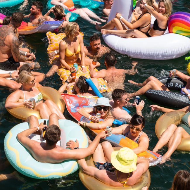 A group of people relaxing and socializing on various inflatable floats in a body of water on a sunny day; many are holding beverages, enjoying a private yacht charter in Sydney Harbour.