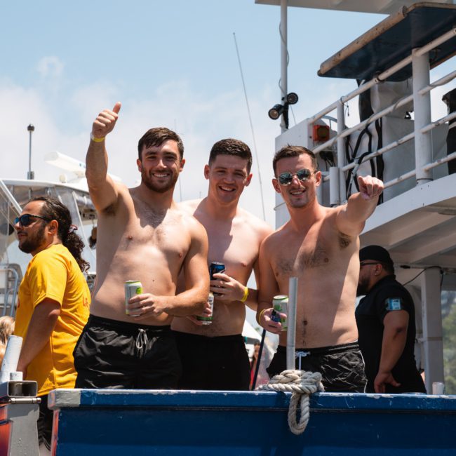 Three shirtless men on a boat are smiling and giving a thumbs-up gesture, enjoying a lively Sydney boat party hire. Other people and boats are visible in the background.