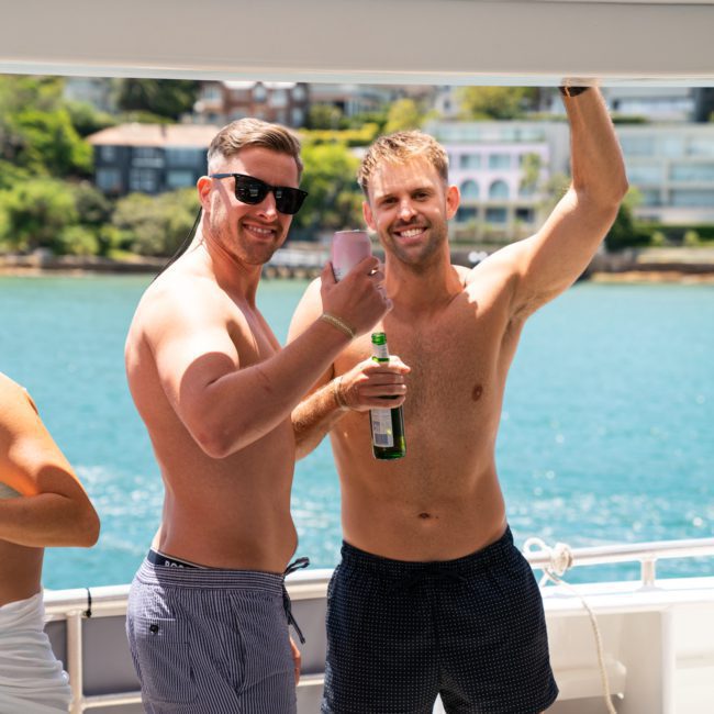 Two men wearing sunglasses and swim shorts stand on a boat deck, smiling and raising drinks, with waterfront buildings in the background—an ideal scene for a private yacht charter on Sydney Harbour.