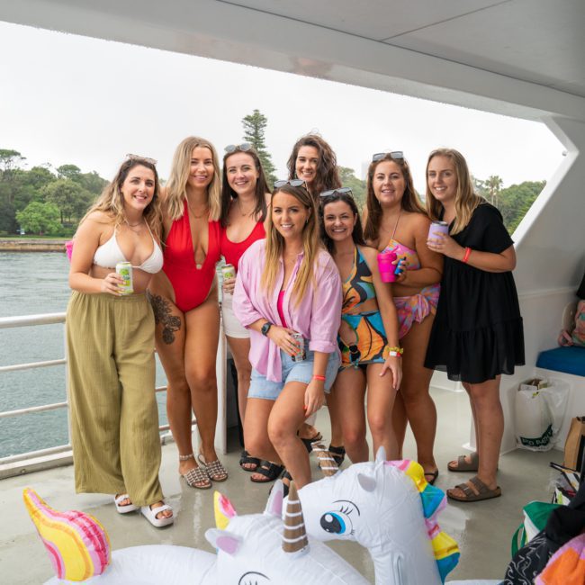 A group of eight women, some holding drinks, pose together on a boat deck with water and trees in the background. Inflatable pool toys are visible in the foreground, capturing the essence of a luxury yacht hire Sydney experience.