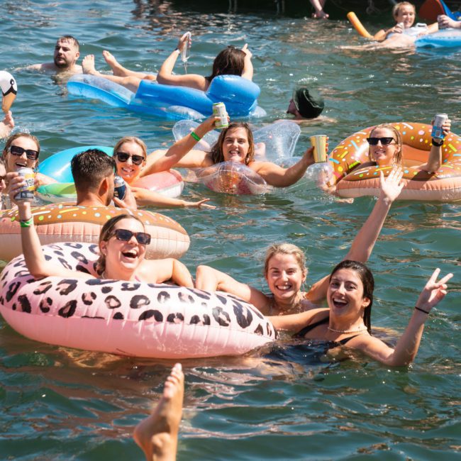 A group of people enjoying a sunny day in the water with inflatable rings and drinks, smiling and waving at the camera during a lively Sydney boat party hire.
