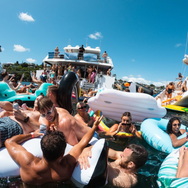 A vibrant scene of people in swimsuits enjoying a sunny day on inflatable loungers and pool floats in the water beside docked boats, perfect for a private yacht charter Sydney Harbour experience.