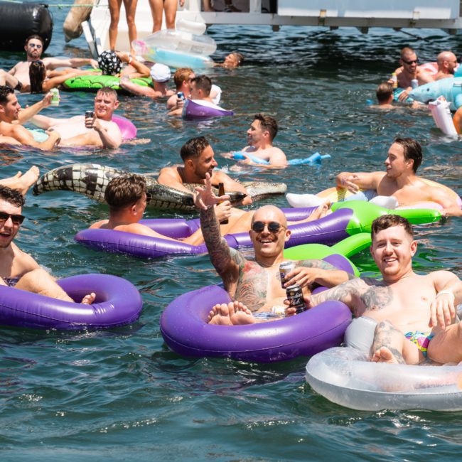 A group of people float on inflatable tubes in a lake, enjoying the sunny weather. Some are holding drinks and engaged in conversation, while a luxury yacht hire Sydney is visible in the background.