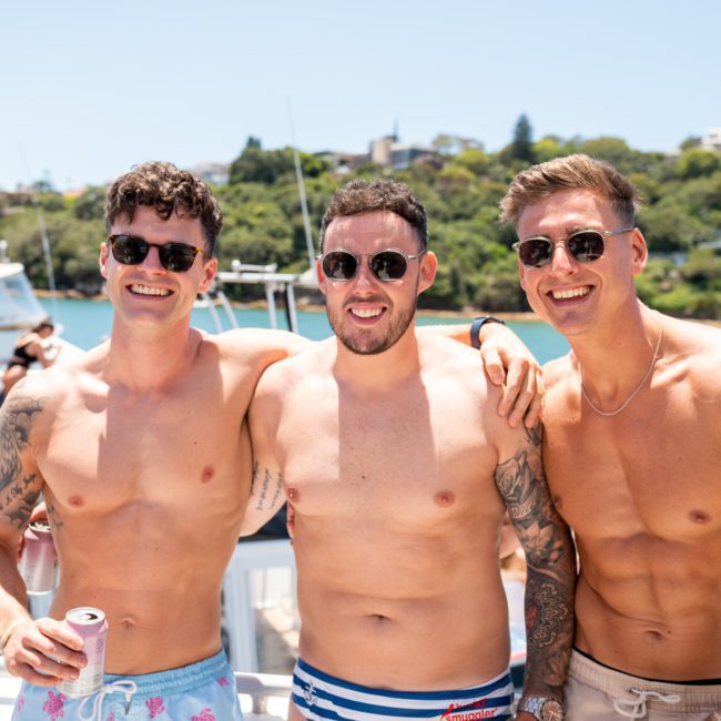 Three men in swim trunks and sunglasses stand together smiling on a boat during a Sydney boat party hire. Other people and scenic waterfront buildings are visible in the background.