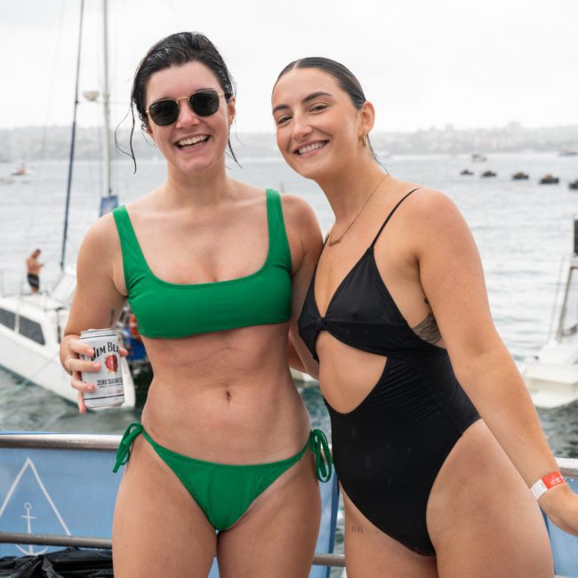 Two people in swimsuits, one in green and one in black, smiling at the camera. One is holding a beverage can. They are standing outdoors near a body of water with boats in the background, enjoying a Sydney boat party hire.