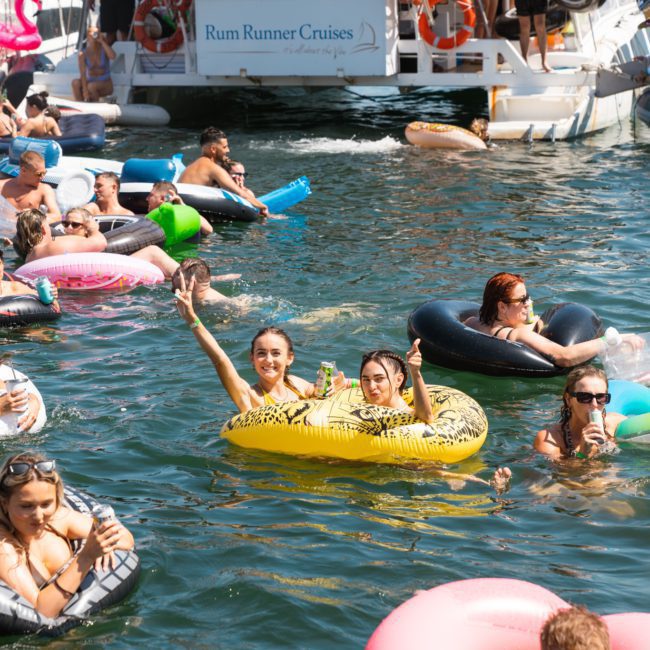 People relax on inflatables in the water near a boat with a "Rum Runner Cruises" sign. Some are waving and socializing under sunny conditions, enjoying the ambiance of a Catamaran party Sydney.