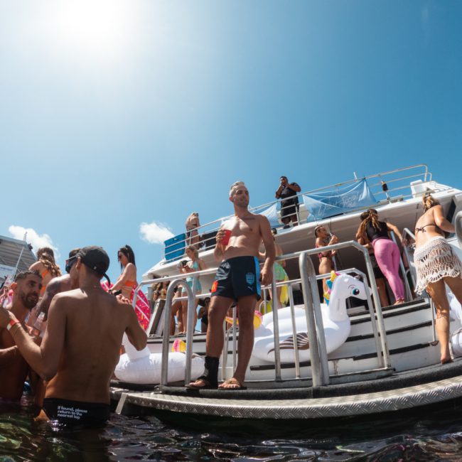 A group of people are gathered on the deck of a white boat, some wearing swimwear, with floaties in the water. The sky is clear with a few clouds. It's perfect for a Catamaran party in Sydney.