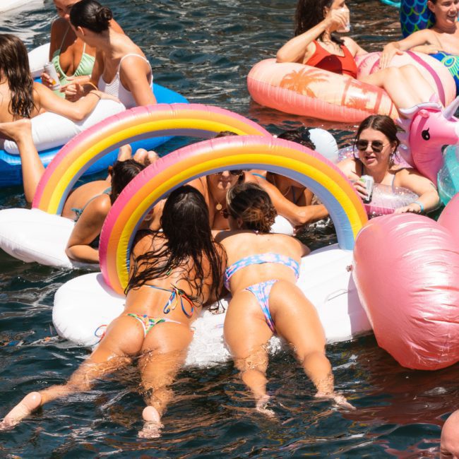 A group of people lounging on various pool floats in the water, including rainbow and flamingo-shaped inflatables, enjoying a sunny day. Some hold drinks while others converse and relax, reminiscent of the fun atmosphere found at luxury yacht hire Sydney events.