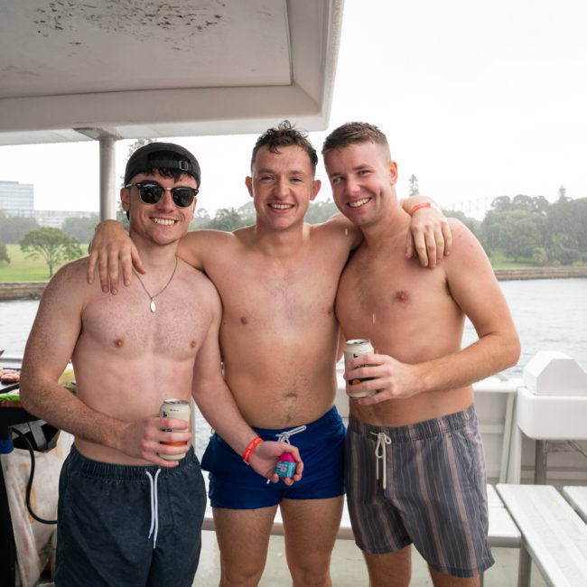 Three shirtless men on a boat smile and pose for a photo, each holding a beverage can. The background shows a waterfront scene with green trees and overcast skies, epitomizing the fun atmosphere of luxury yacht hire Sydney.