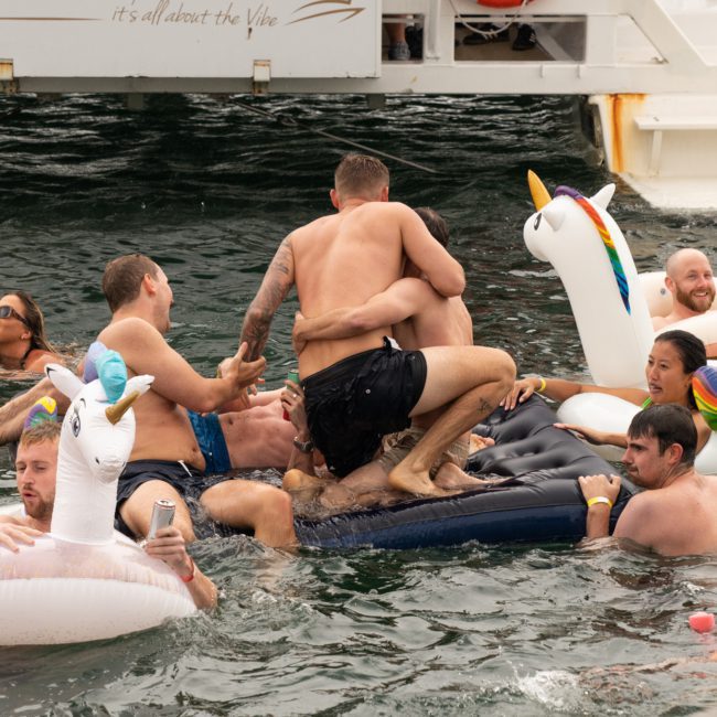 A group of people in swimwear are enjoying a Catamaran party Sydney in a body of water with inflatable toys and unicorn floaties. They are in front of a boat, some standing, sitting, or splashing around.