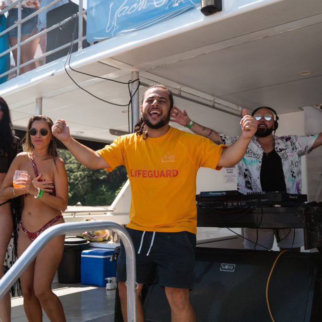 A man in a yellow "Lifeguard" shirt and black shorts is smiling and raising his arms on a boat. A DJ and several guests are in the background, enjoying the "Yacht Social Club" event.