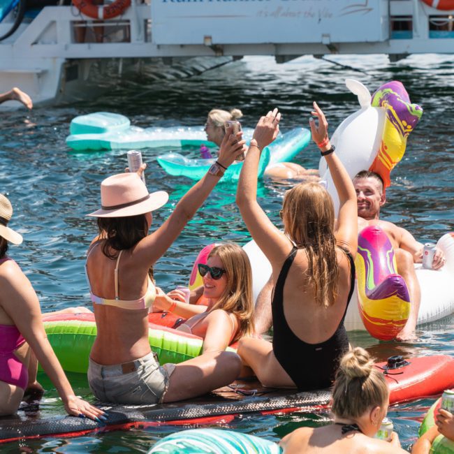 A group of people enjoy a sunny day on colorful inflatables in the water, with a "Rum Runner Cruises" boat in the background. They are laughing and holding drinks up in celebration at a vibrant Sydney boat party hire event.
