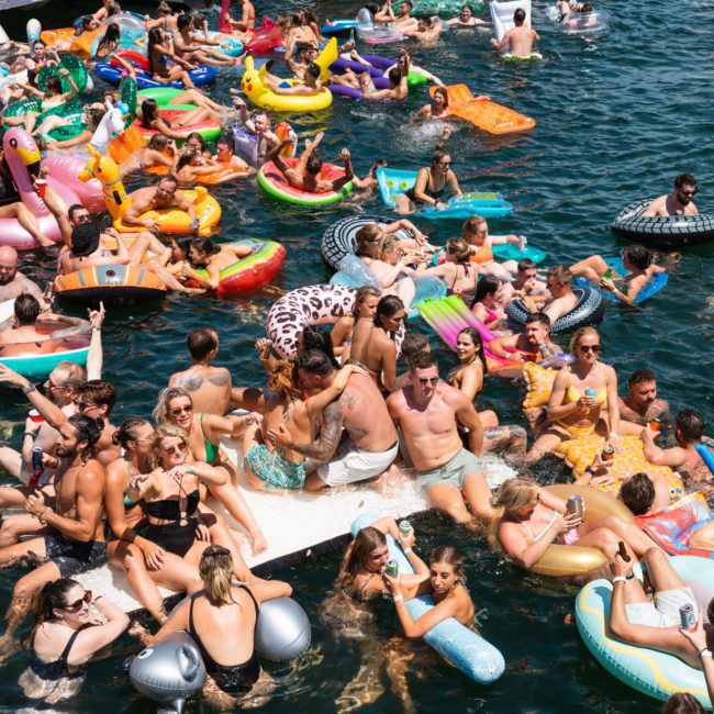 A large group of people on inflatable tubes and rafts float on the water, forming a vibrant and crowded scene at a Sydney boat party hire event.