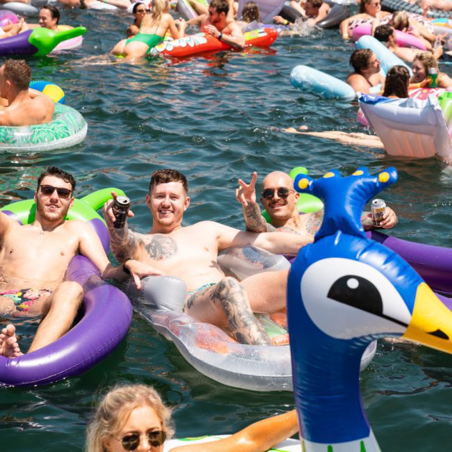 People float on inflatable pool toys in a crowded body of water on a sunny day, with smiles and drinks in hand, as a DJ boat hire Sydney sets the perfect party vibe.