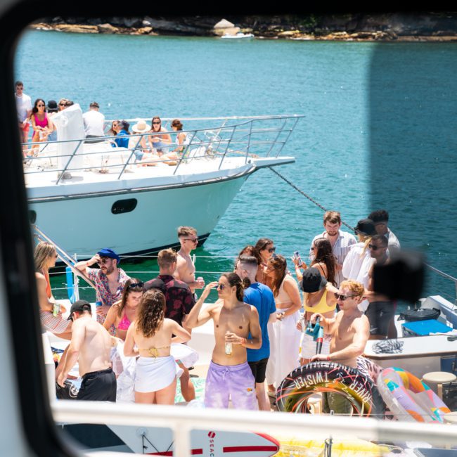 A group of people in swimwear are gathered on a boat, socializing and having fun near another boat docked in a clear blue water area. The scene, viewed from the window of a nearby vessel, captures the vibrant energy of a Catamaran party Sydney.