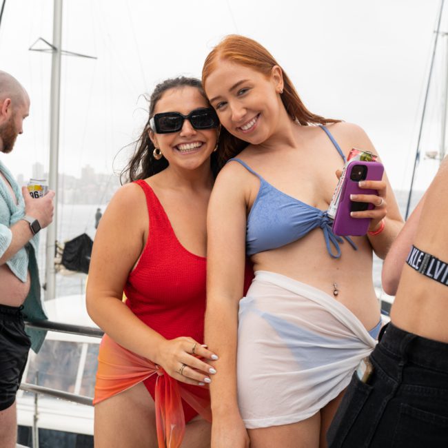 Two women, one in a red swimsuit and the other in a blue swimsuit, stand together on a luxury yacht hire in Sydney, smiling and holding phones. In the background, additional people and parts of the boat are visible.