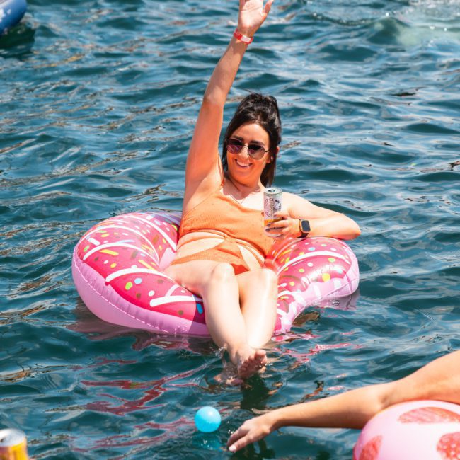 A person floats on a pink inflatable in a body of water, raising one arm and holding a beverage in the other hand, surrounded by other people and inflatables during an unforgettable catamaran party in Sydney.