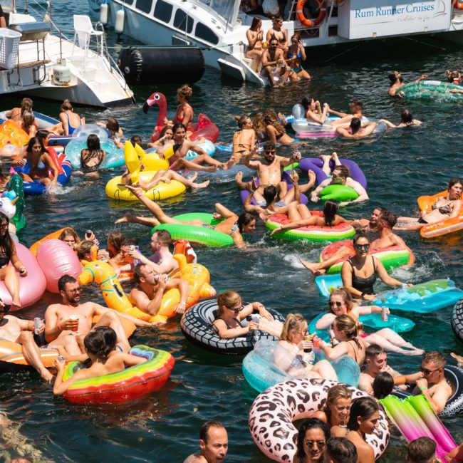 A large group of people on inflatables enjoy a catamaran party in Sydney near boats. The scene includes colorful floaties with a backdrop of clear blue water and a sunny sky.