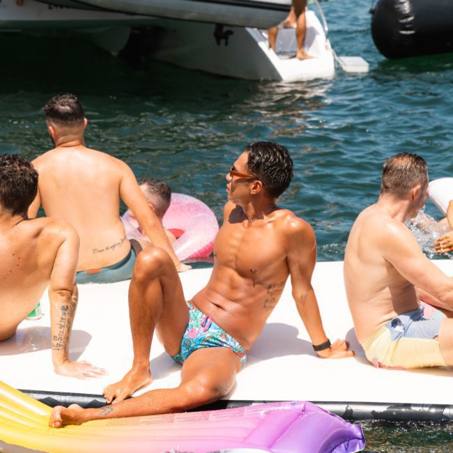 A group of individuals enjoying a sunny day on a lake. One person is sitting on a rainbow-colored float while others are gathered on an inflatable platform near a boat, reminiscent of the vibrant atmosphere of a Catamaran party in Sydney.