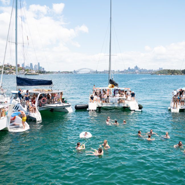 People on several anchored boats and in the water enjoying a sunny day with a city skyline in the background. Some people are swimming while others are on inflatable rafts, making it the perfect setting for a Sydney boat party hire or corporate boat events Sydney.