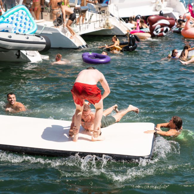 People enjoying a sunny day on a lake, with some lounging on luxury yacht hires and others swimming or playing on inflatables in the water.