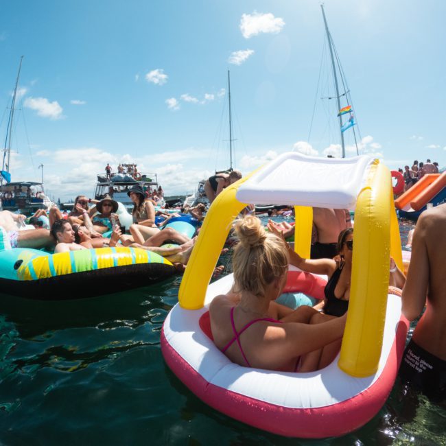 People are leisurely floating on inflatable rafts and boats in the water on a sunny day, creating a festive atmosphere reminiscent of a DJ boat hire Sydney event.