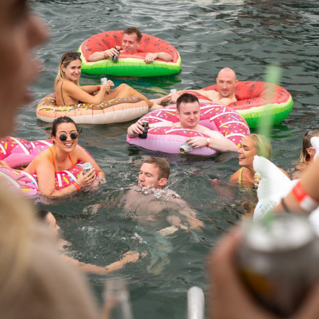People enjoying a swim and floating on inflatable pool tubes shaped like donuts and watermelons in the water near a luxury yacht hire Sydney.