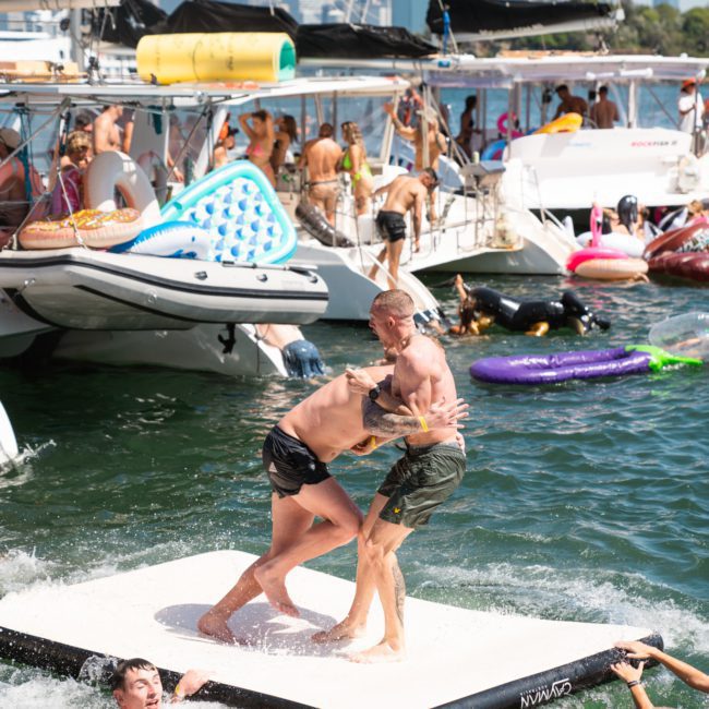 Two men are playfully wrestling on a floating mat in the water while others enjoy the scene from nearby boats and swim around, making it a perfect moment for those considering Sydney boat party hire.