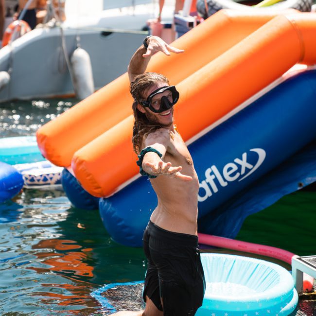 A person wearing swim goggles and a snorkel poses playfully in the water, with inflatable structures and a boat hosting a DJ boat hire Sydney event in the background.