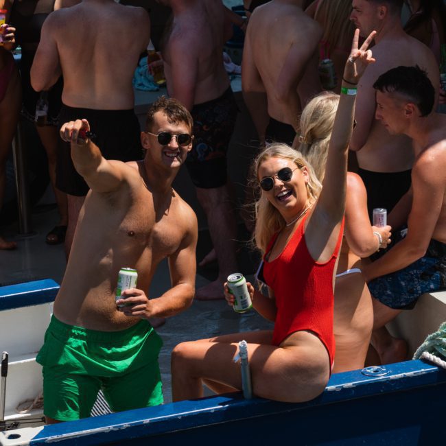 People enjoying a catamaran party in Sydney, with a man in green shorts and a woman in a red swimsuit smiling and gesturing towards the camera. Various guests in swimwear are mingling in the background.