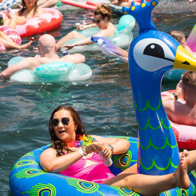 People enjoying a sunny day on a lake, floating on colorful inflatables including one shaped like a peacock, reminiscent of the fun had at Sydney boat party hire events.