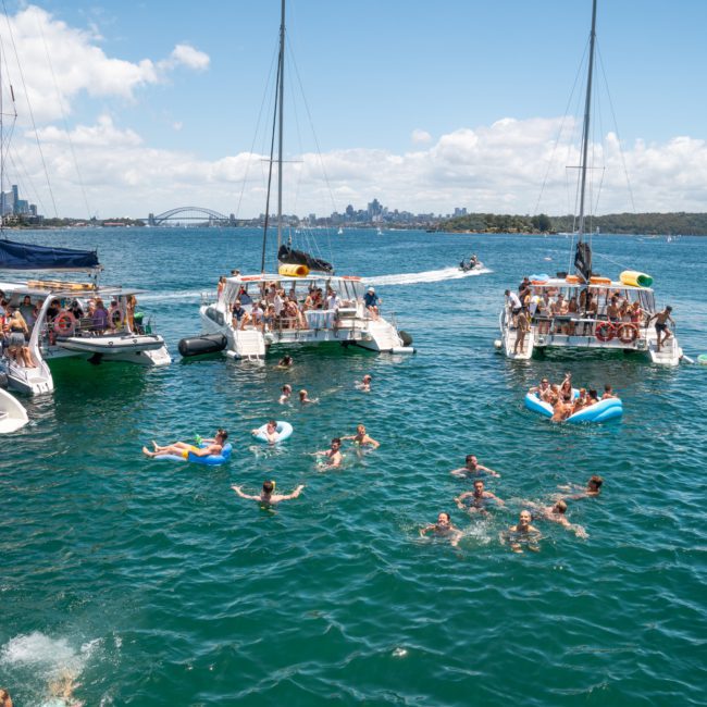 A group of people swim and float on inflatables in a body of water, surrounded by three boats. The city skyline and a bridge are visible in the background under a sunny sky, perfect for a catamaran party Sydney.