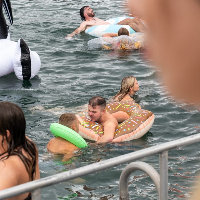 People are swimming and floating on inflatable pool toys in a body of water, with an inflatable penguin visible. Some individuals are interacting near a railing, enjoying the lively atmosphere often found at Corporate boat events Sydney.