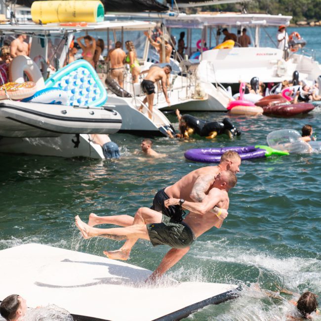 Two men playfully wrestle and fall into the water from a floating mat among a group of people swimming near boats anchored by the shore, with a city skyline visible in the background. It looks like everyone is having an incredible time on their Catamaran party Sydney.