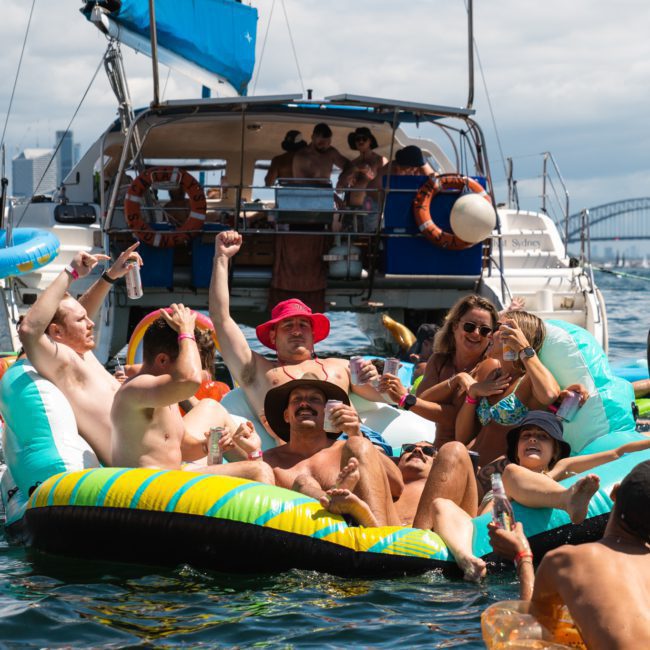 A group of people floats on inflatable rafts near a boat in a body of water, enjoying a lively party. The Sydney skyline and Harbour Bridge are visible in the background, showcasing the perfect setting for a Sydney boat party hire.