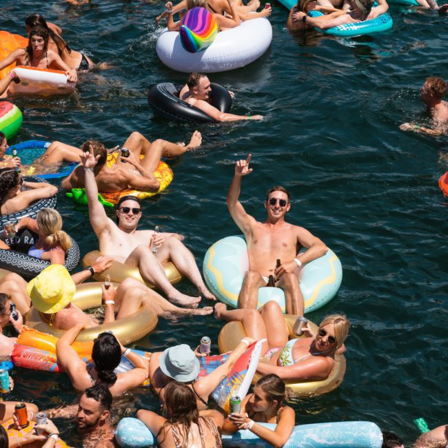 A group of people on inflatable floats and swim rings relax and socialize in a crowded body of water under the sun, some holding drinks, while others admire a private yacht charter in Sydney Harbour nearby.