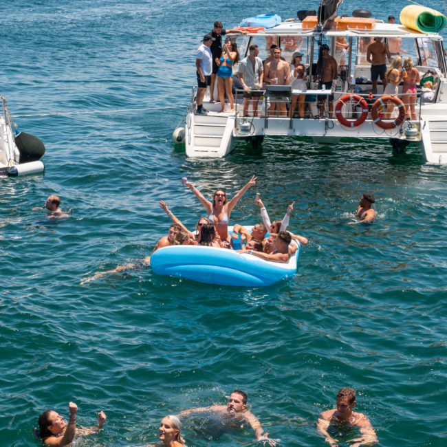 People swimming and relaxing on inflatable floats in the water near two boats on a sunny day, enjoying a Catamaran party in Sydney.