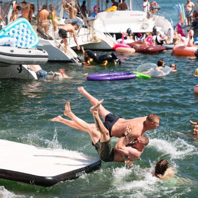 People are enjoying a day on the water, with some jumping from boats and floating on inflatables. Three people are playfully falling into the water, while others swim and relax nearby during a Catamaran party in Sydney.