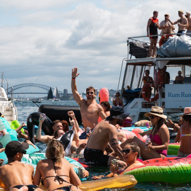 A group of people on inflatable floats enjoy a sunny day on the water near anchored boats. One man waves towards the camera while others relax and socialize. In the background, a bridge is visible, setting the scene for what could be an amazing private yacht charter Sydney Harbour experience.