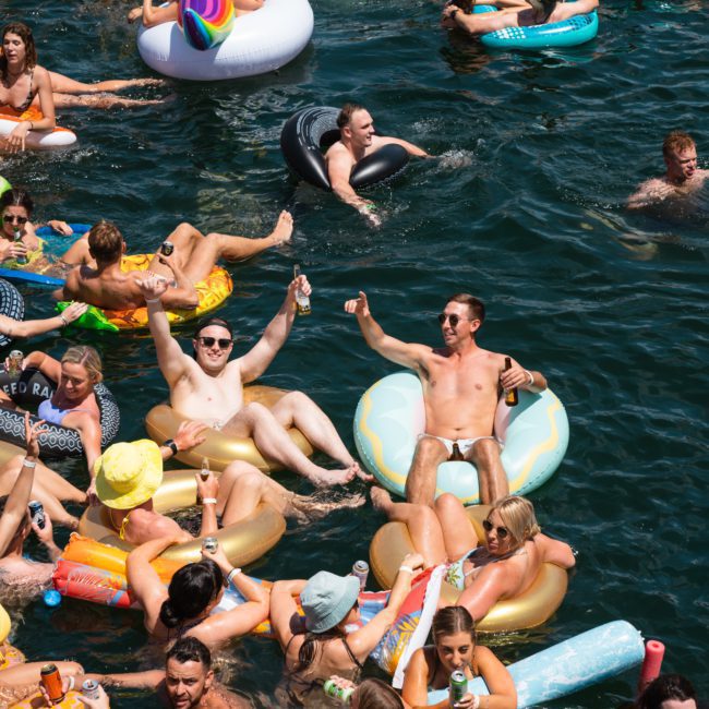 A lively group of people in inflatable pool floats are enjoying themselves in the water, holding drinks and socializing on a sunny day, reminiscent of a Catamaran party Sydney.