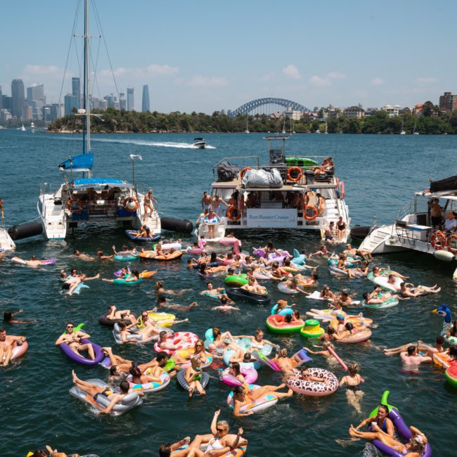 A group of people are relaxing on colorful inflatable floaties in the water between several boats on a sunny day, with a city skyline and bridge visible in the background, enjoying what looks like a luxurious Sydney boat party hire.