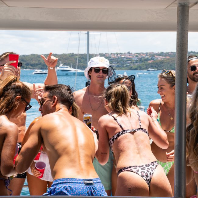 A group of people in swimwear are socializing and dancing on a luxury yacht hire in Sydney with a body of water and another boat visible in the background. Some people hold red cups.