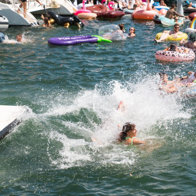 People swimming and using flotation devices in a crowded body of water near multiple boats on a sunny day, with a luxury yacht hire in Sydney adding to the vibrant atmosphere.