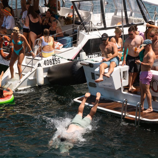 A group of people enjoy a sunny day on two boats docked side by side. One person is diving into the water while others relax on the luxurious yachts, socializing and watching the diver.