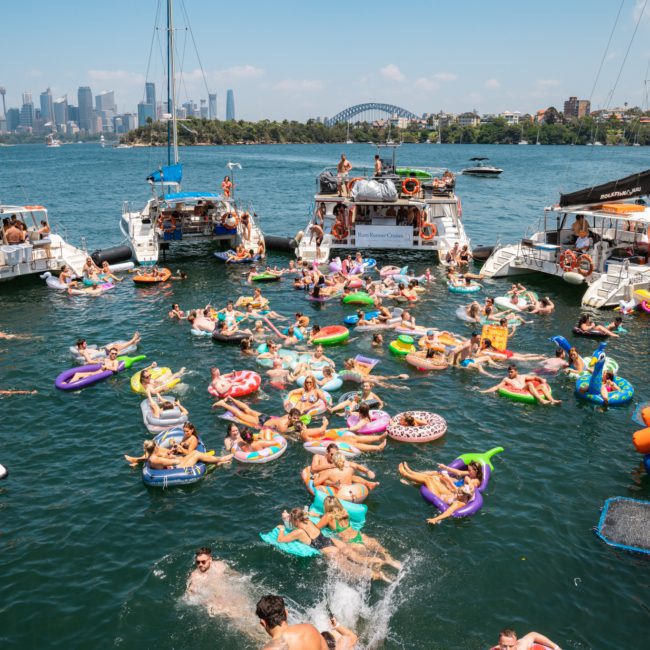 A large group of people are gathered on inflatable pool floats and boats in a bay, with a city skyline and bridge visible in the background, perfect for a Sydney boat party hire or private yacht charter Sydney Harbour.