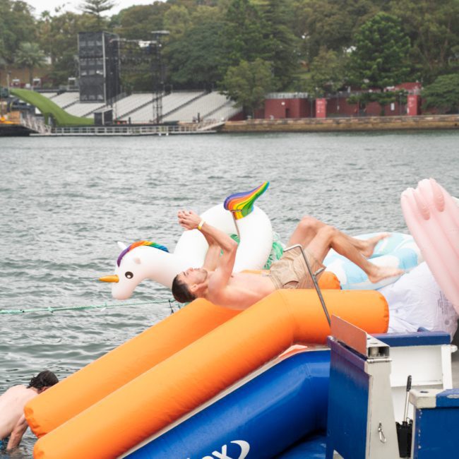 A person is lying on a large inflatable unicorn float in the water near a dock. The scene appears to be at a recreational waterfront area with trees and an amphitheater in the background, reminiscent of a Catamaran party Sydney.