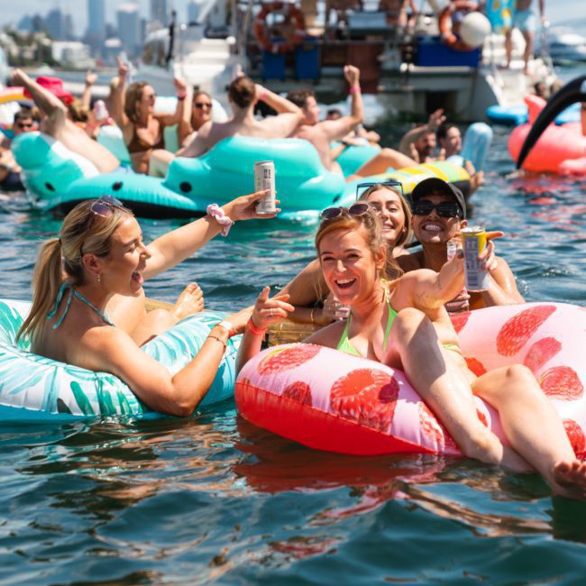 A group of people on inflatable pool floats enjoy drinks and socialize on a sunny day, surrounded by other floats and boats on the water during a Sydney boat party hire.
