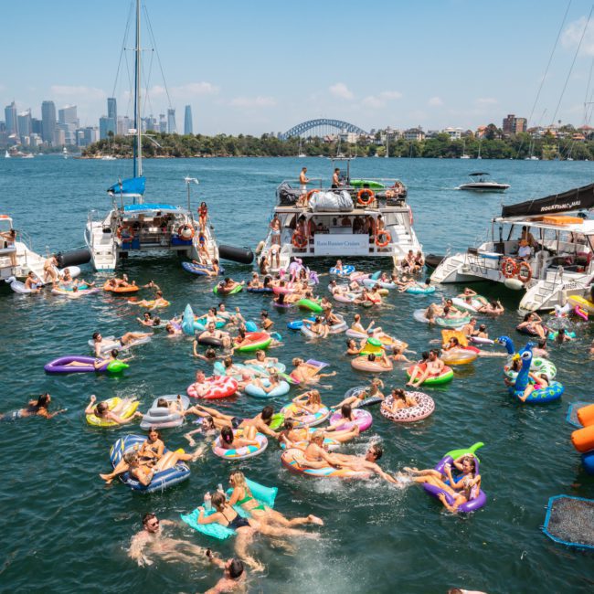 People are gathered on inflatable floats and boats in a busy waterway, enjoying the vibrant atmosphere with a city skyline and bridge in the background. Nearby, a private yacht charter Sydney Harbour glides smoothly across the water.